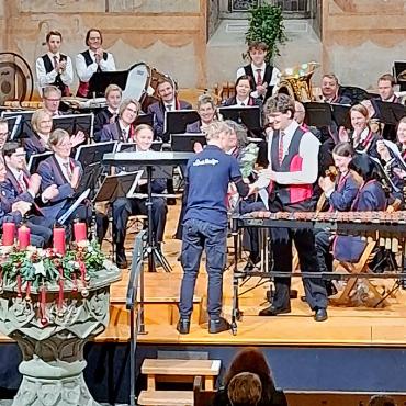 Klangvolle Einstimmung auf die Adventszeit - Nils Romang wird von seinen Musikkolleg:innen, dem Dirigenten Sandro Romang (links stehend) und allen Anwesenden für seinen tollen Auftritt am Xylofon bejubelt und erhält ein Geschenküberreicht von einem Junior-Band-Mitglied.