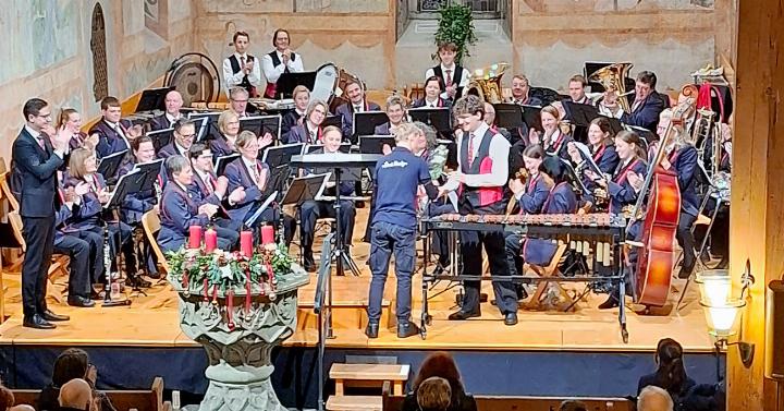 Nils Romang wird von seinen Musikkolleg:innen, dem Dirigenten Sandro Romang (links stehend) und allen Anwesenden für seinen tollen Auftritt am Xylofon bejubelt und erhält ein Geschenküberreicht von einem Junior-Band-Mitglied.